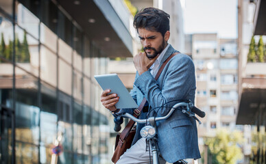 Young worried businessman using digital tablet outdoors while commuting with bicycle
