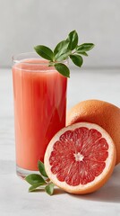 Freshly Squeezed Pink Grapefruit Juice in a Tall Glass with Whole and Halved Grapefruits and Green Leaves on a Textured Light Gray Surface with Soft Natural Lighting
