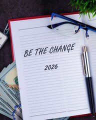 On a desk, the phrase Be the change stands out on a notepad, surrounded by cash, a pen, glasses, and a small plant, symbolizing hope for 2026 and personal growth