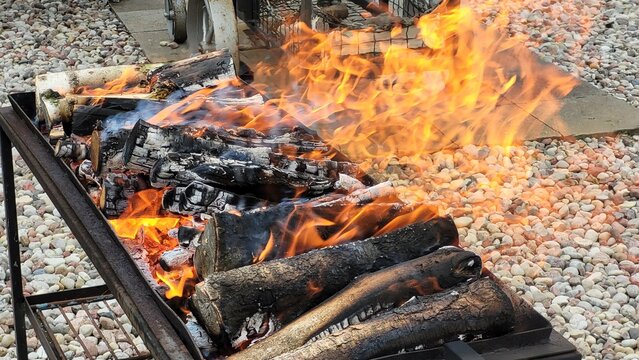 Burning firewood in a metal grill. Bright orange flames and glowing embers rise above blackened logs, surrounded by a stone-covered yard.