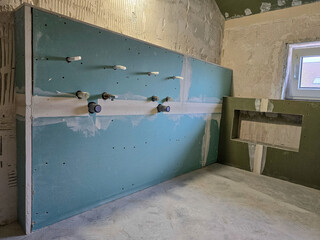 bathroom during renovation shows green drywall with visible plumbing outlets and taped seams, ready for further installation under natural light from a small window