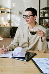 Middle aged Caucasian woman wearing glasses sitting at desk holding cup of coffee while writing in notebook, working in modern office environment with documents on table