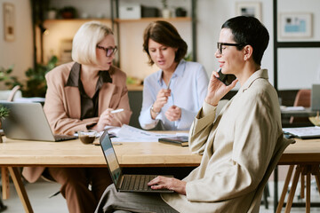 Middle aged Caucasian woman sitting at desk using laptop and talking on smartphone, while two middle aged Caucasian women discussing documents in background during business meeting