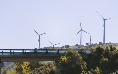 Row of modern wind turbines on green hillside behind highway bridge, clean renewable energy landscape under clear blue sky, sustainable electricity production concept
