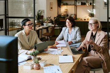Three middle aged Caucasian women sitting at table discussing business strategy, using laptop and...