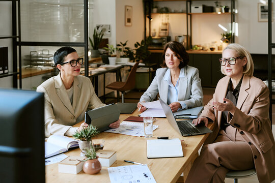Three middle aged Caucasian women sitting at table discussing business strategy, using laptop and digital tablet, taking notes during professional meeting in modern office workspace