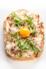 A creative, delicious flatbread topped with prosciutto, fresh arugula, shredded cheese, and a raw egg yolk ready to be mixed, captured from above on a white background.