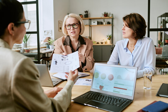Three middle aged Caucasian women discussing business analytics at table, reviewing printed charts and graphs, using laptops, collaborating on financial strategy in modern office