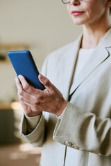 Caucasian middle aged woman using smartphone, holding device with both hands, standing indoors, lips visible but eyes out of frame, wearing business attire, focused on screen