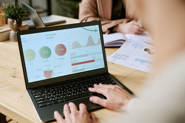 Caucasian young adult man working on laptop analyzing business data charts while sitting at desk in office, another Caucasian young adult woman reviewing documents in background