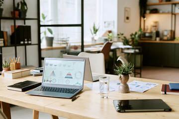 Modern office workspace showing open laptop with business charts on screen, digital tablet,...