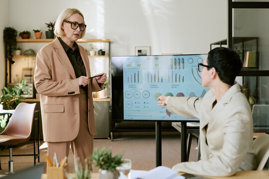 Caucasian middle aged woman presenting business data on digital screen while Caucasian young adult woman sitting and pointing at chart during office meeting