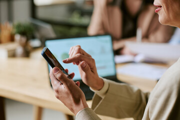 Caucasian middle aged woman using smartphone while sitting at desk, holding device in one hand and...