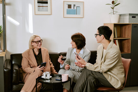 Three middle aged Caucasian women sitting together on sofa engaging in conversation, holding smartphones and drinking coffee, smiling and interacting in modern office lounge