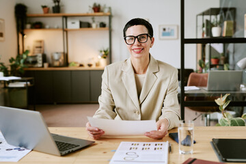 Portrait of middle aged Caucasian woman with short hair wearing glasses sitting at desk holding...