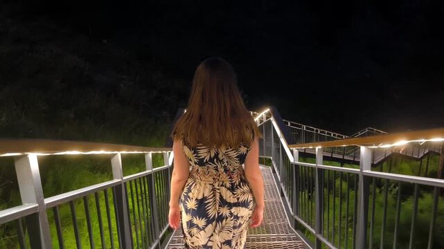 A woman in a summer dress walks down a lighted staircase in the dark. Romantic evening walk, hiking and recreation. Symmetrical perspective, no face.
