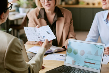 Three middle aged Caucasian women discussing business analytics, one handing printed charts to...