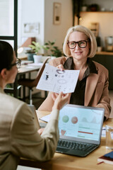 Caucasian middle aged woman wearing glasses sitting at desk discussing business charts with...