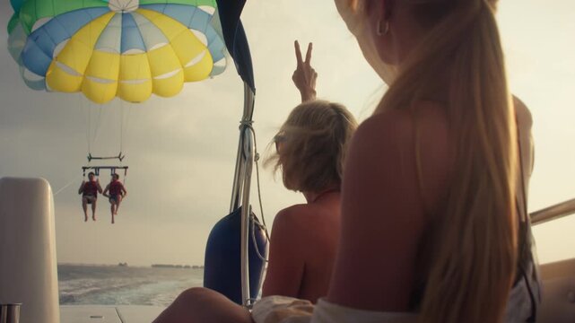 Friends observe two people parasailing high above the water from a boat. The vibrant parachute adds color to the serene sky, creating a memorable vacation moment.