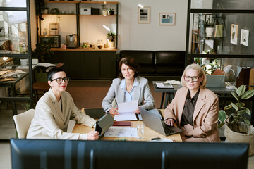 Portrait of three middle aged Caucasian women sitting at desk working with laptops and documents,...
