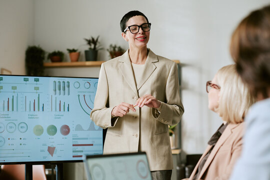 Caucasian middle aged woman presenting business data to group of middle aged and senior women in office setting, standing near digital display with charts and graphs, engaging audience