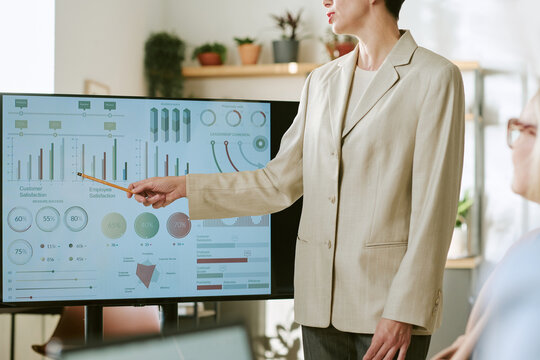 Caucasian middle aged woman presenting business analytics on large screen to colleague, using pointer to highlight customer and employee satisfaction charts during meeting - Powered by Adobe
