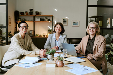 Three middle aged Caucasian women sitting at conference table discussing business strategy, smiling...