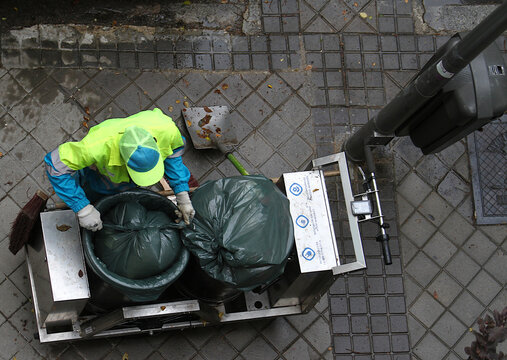 Barrendero basurero de mantenimiento de limpieza atando y anudando una bolsa de basura y residuos en cubo con carro y papelera en acera de calle del ayuntamiento de Madrid desde arriba en perspectiva