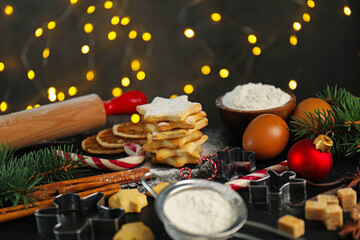 Christmas composition with cookies in shape of stars, cutters, ingredients and fir tree branches on black table against blurred lights, closeup