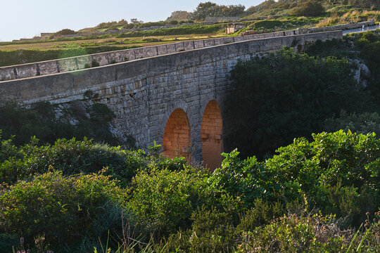 Historic stone bridge along Victoria Lines in Malta, glowing arches at sunset, green valley and shrubs in foreground