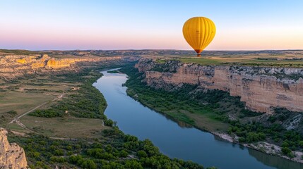 A yellow hot air balloon floats serenely over a green canyon with a winding river below, captured in soft afternoon light
