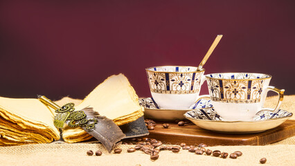 A beautifully composed still-life image of two elegant tea cups with golden patterns, placed beside a vintage quill and stack of paper, with coffee beans scattered on a burlap surface. Ideal for use 