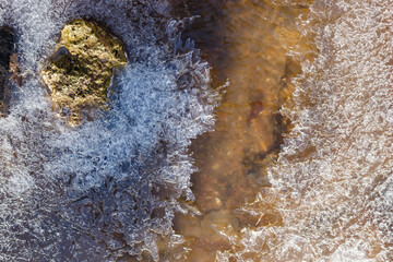 Intricate ice crystals adorn a cold stream, partially covering a mossy rock and clear, flowing water. A beautiful natural winter texture