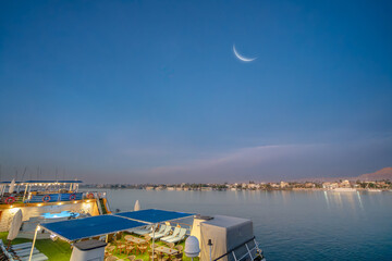 Springtime dawn view of a crescent moon above Luxor West Bank in Egypt, taken from the deck of a peaceful Nile River cruise ship with lounge chairs.