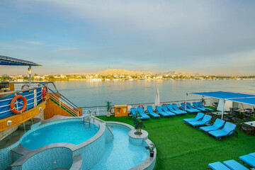 Springtime dawn view from a cruise ship pool and sun deck with lounge chairs, overlooking the Nile River and the scenic Luxor West Bank in Egypt.