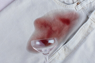 Spilled glass with red wine falled a top a light blue pair of jeans, while a noticeable red stain spreads from the glass. Daily stains concept. top view