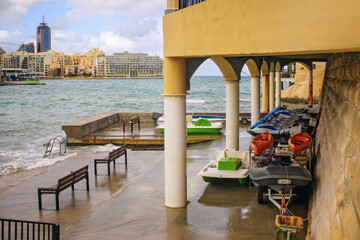Boats and jet skis under arched shelter on a wet pier with benches against stormy Mediterranean Sea...