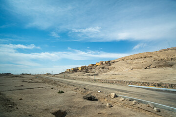 Wide low angle view of a highway passing through the arid desert landscape near the Colossi of Memnon Luxor Egypt with small traditional buildings on a distant hill.