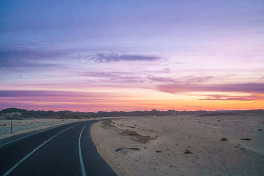 The desert highway curves through rocky, sandy terrain under a breathtaking twilight sky with vibrant pink and purple clouds on the road to Hurghada Red Sea resort Egypt.