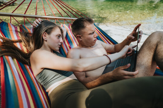 A young couple relaxes in a colorful hammock by the water while working remotely on their laptop, combining leisure and productivity in a serene natural setting.