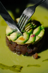 Close-up of a gourmet tuna tartare with avocado slices and edible flowers, being cut with a fork and knife on a green sauce plate. Elegant fine dining concept.

