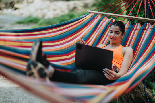 A woman using a laptop while relaxing on a colorful striped hammock outdoors on a sunny day. - Powered by Adobe