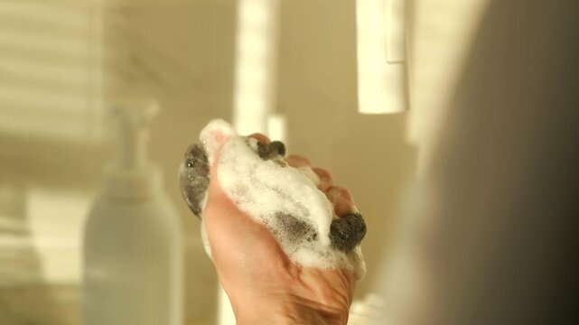 Close up view of a hand squeezing a dark gray sponge, creating white sudsy foam from dish soap in a kitchen sink with warm morning light streaming through a window in the background