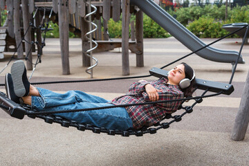 Young woman in a plaid shirt and headphones relaxing on a rope swing in a modern residential...