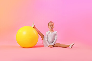 Smiling little girl with fitness ball doing gymnastic exercise on pink gradient background
