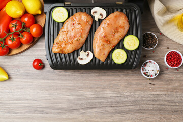 Electric grill with raw marinated chicken fillet, mushrooms, spices and products on wooden table, flat lay. Space for text