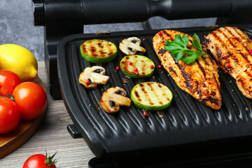 Electric grill with tasty chicken fillet, mushrooms, products and spices on wooden table against grey background, closeup