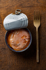 Open tin can of salmon fish fillet in its own juice and metal fork on wooden table background, closeup, top view