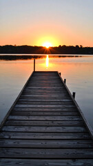 Fototapeta premium Wooden pier extending into calm water during sunrise with orange sky