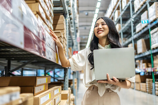 Engaging in meticulous inventory management, a young asian woman professional examines products while using a laptop computer, surrounded by organized shelves filled with boxes